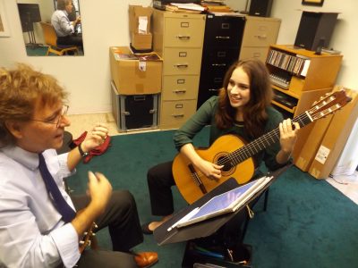 The student and professor are seated side by side in his teaching studio as he simulates playing the guitar.