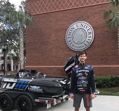 Stetson MBA student Thomas Oltorik stands in his fishing tournament clothes and beside his bass speed boat in front of the Rinker Welcome Center.