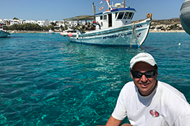 Jason Maddox poses at the water's edge with a fishing boat in the background and crystal clear blue water.