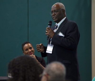 Jim Johnson stands in the front of the crowd with a microphone as Gwen Azama-Edwards listens on the panel