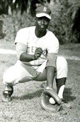 Jim Johnson wears Stetson baseball uniform in B&W photo, reaches down and has ball in his catcher's mitt.