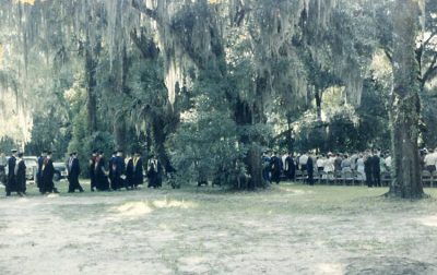 A processional of professors and administrators walked into a wooded area with chairs in this black and white archival photo