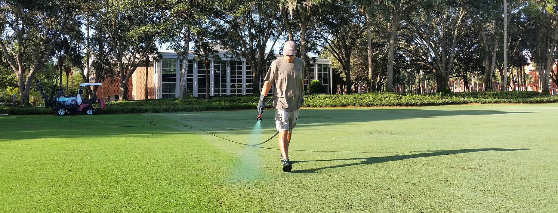 wide shot of Steve spraying a green liquid on the grass with a long hose trailing to his workcart. In the background is the duPont-Ball Library.