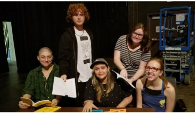 Group shot of all of them sitting and standing around a table with scripts.