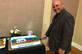 Chief Matusick stands with knife in hand, cutting a sheet cake with the word, retirement, and a plastic motorhome on top.
