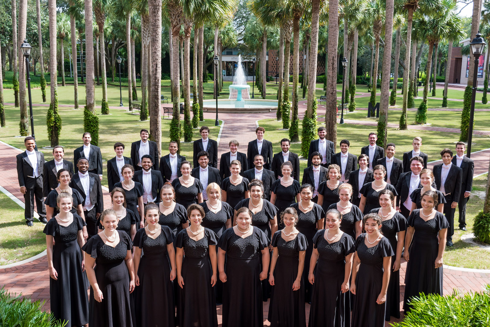 Big group shot of the entire choir in black tuxes and black gowns standing in front of the fountain in Palm Court.