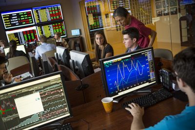 Students work at computers in a stock trading room as a professor looks on