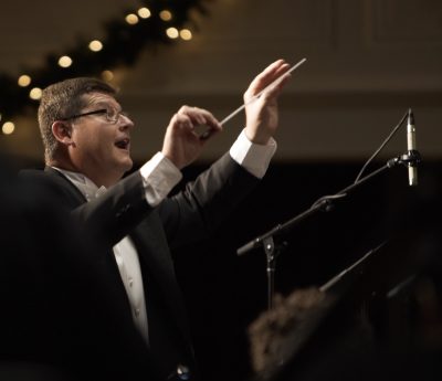 Timothy Peter in tuxedo holds baton mid-air as he conducts