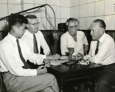 A black and white photo of a student and three older men around a microphone in a cramped room.
