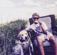 Joseph Cofer is sitting in a chair along the dunes of the beach with a yellow labrador retriever by his side.