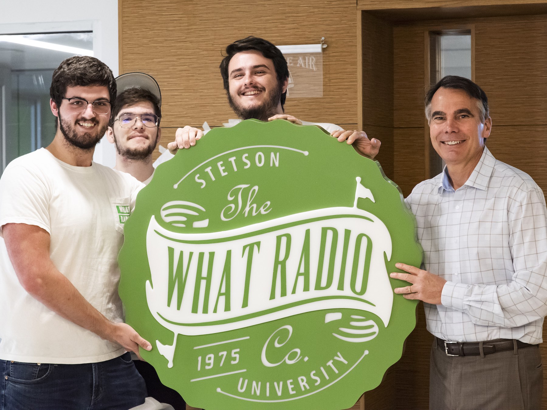 The four men hold a green and white sign that says WHAT Radio outside the radio studio in the CUB