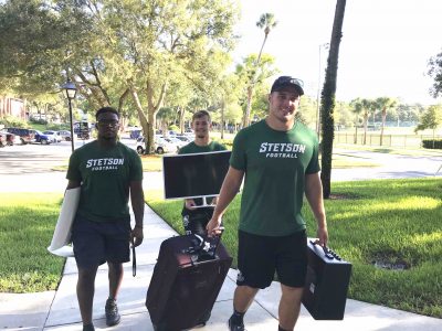 The three players come up a walkway carrying items into a residence hall.