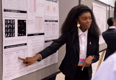 Heyley Gatewood stands at a display board with math and science displays, talking to attendees at a conference.