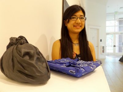 Student stands in art gallery with two pieces, each wrapped in cloth -- one black and one blue