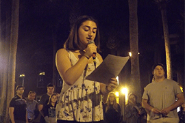 A student holds a microphone and reads from notes in front of a small crowd at night
