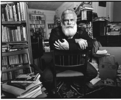 B&W portrait of him seated in an office with lot of books on shelves.