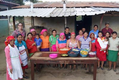 Rebecca Williams stands in a Honduras village with locals, outside a simple wooden shack and a table with bowls of food.