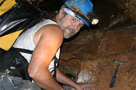 The geologist is in a cave with a lighted helmet and pick axe to get rocks, and a backpack on.