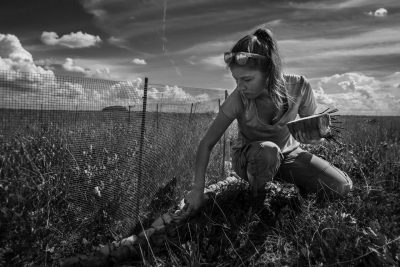 A black and white photo of a woman looking through grassy ground with big sky overhead.