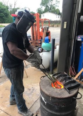 Worker in protective gear puts pieces of metal into fire drum with metal pinchers.