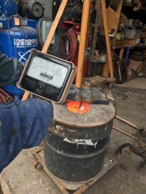 a worker holds a temperate gauge in front of hot cauldron of metal