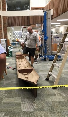The old canoe sits on the carpet in the library with workmen around it.
