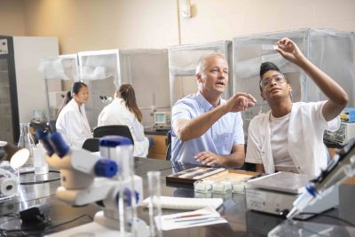 Professor talks to student holding up a microscope slide as two other lab workers look at a microscopic 