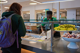 A dining worker dishes food into a to-go container for a waiting student