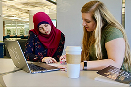 two students are looking at coursework with a laptop open in front of them.