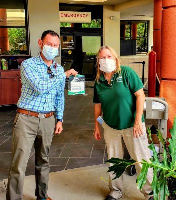 The two of them wear face coverings and he holds up a bag of ear guards outside Hospital ER.