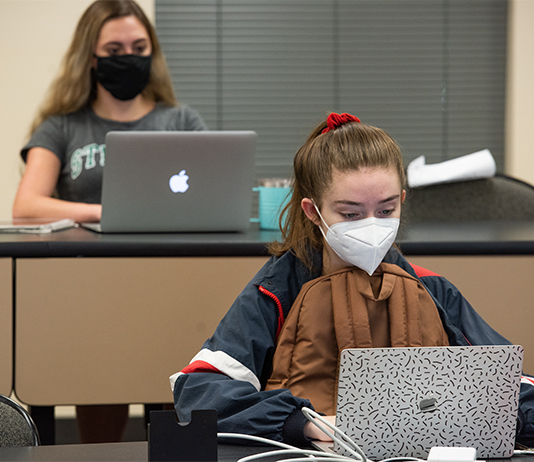 Two students sit in a classroom working on laptops with face coverings on