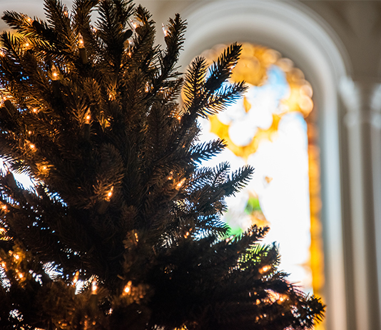 A Christmas tree with white lights and a stained glass Chapel window in the background.