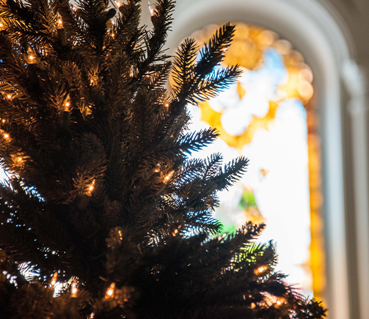 A Christmas tree with white lights and a stained glass Chapel window in the background.