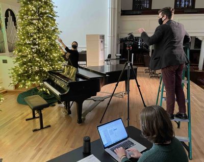 A stage crew set up a camera in front of a piano and a Christmas tree in Lee Chapel