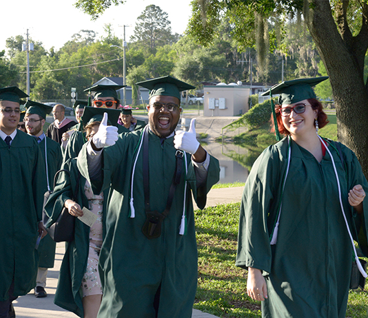 A graduate in a cap and gown gives a thumbs up while walking toward Commencement with other graduates.