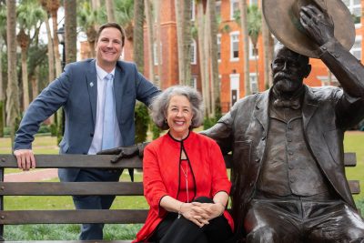 Retired president Wendy Libby sits beside the John B. Stetson statue on campus while current President Christopher Roellke stands behind her.