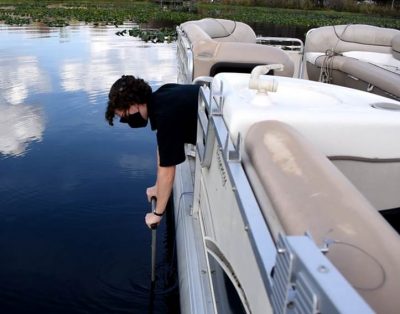 A student leans over the side of a boat to collect a core sample from a lake.