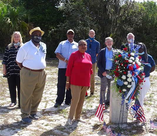 A group of people stand around a grave marker adorned with flags and red and white flowers.