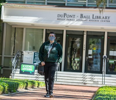 A student walks in front of the library wearing a face mask