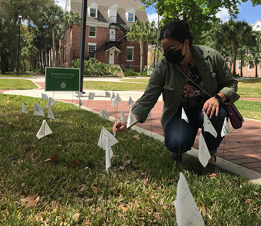 A woman kneels beside a COVID-19 remembrance flag on campus