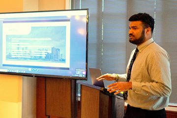 A student stands giving a presentation in front of a digital screen.