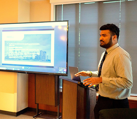 A student stands giving a presentation in front of a digital screen.