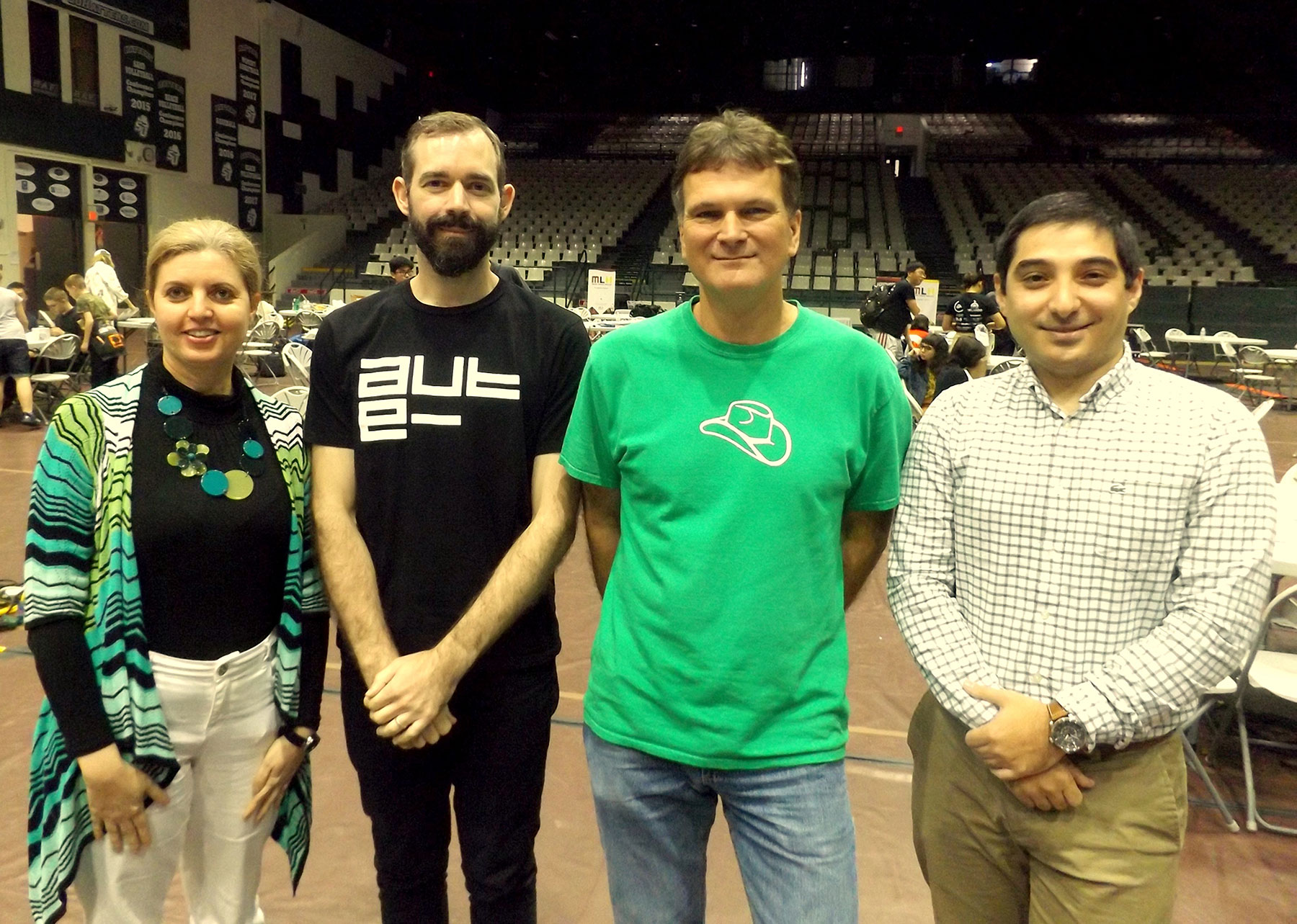 Four professors stand in a busy gymnasium during a hackathon event, including Hala ElAarag