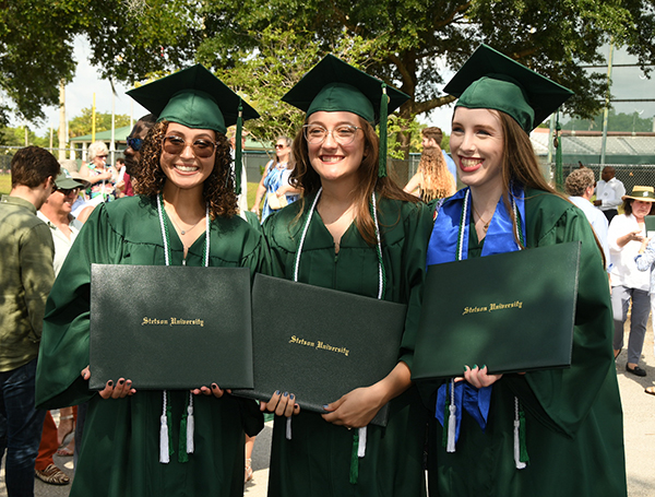 Three women pose in cap and gown, and holding diplomas following Commencement.