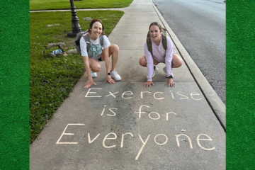 Two students pose by words written in chalk on a sidewalk, "Exercise is for Everyone."