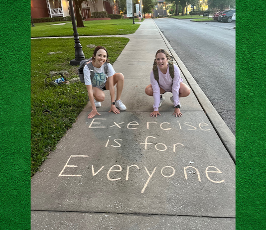 Two students pose by words written in chalk on a sidewalk, "Exercise is for Everyone."
