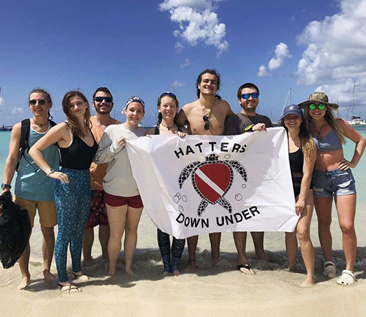 Group shot of students holding a flag that says, Hatters Down Under, on a beach.