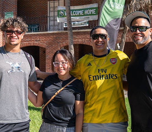 Four people pose in front of a residence hall that says, Welcome Home