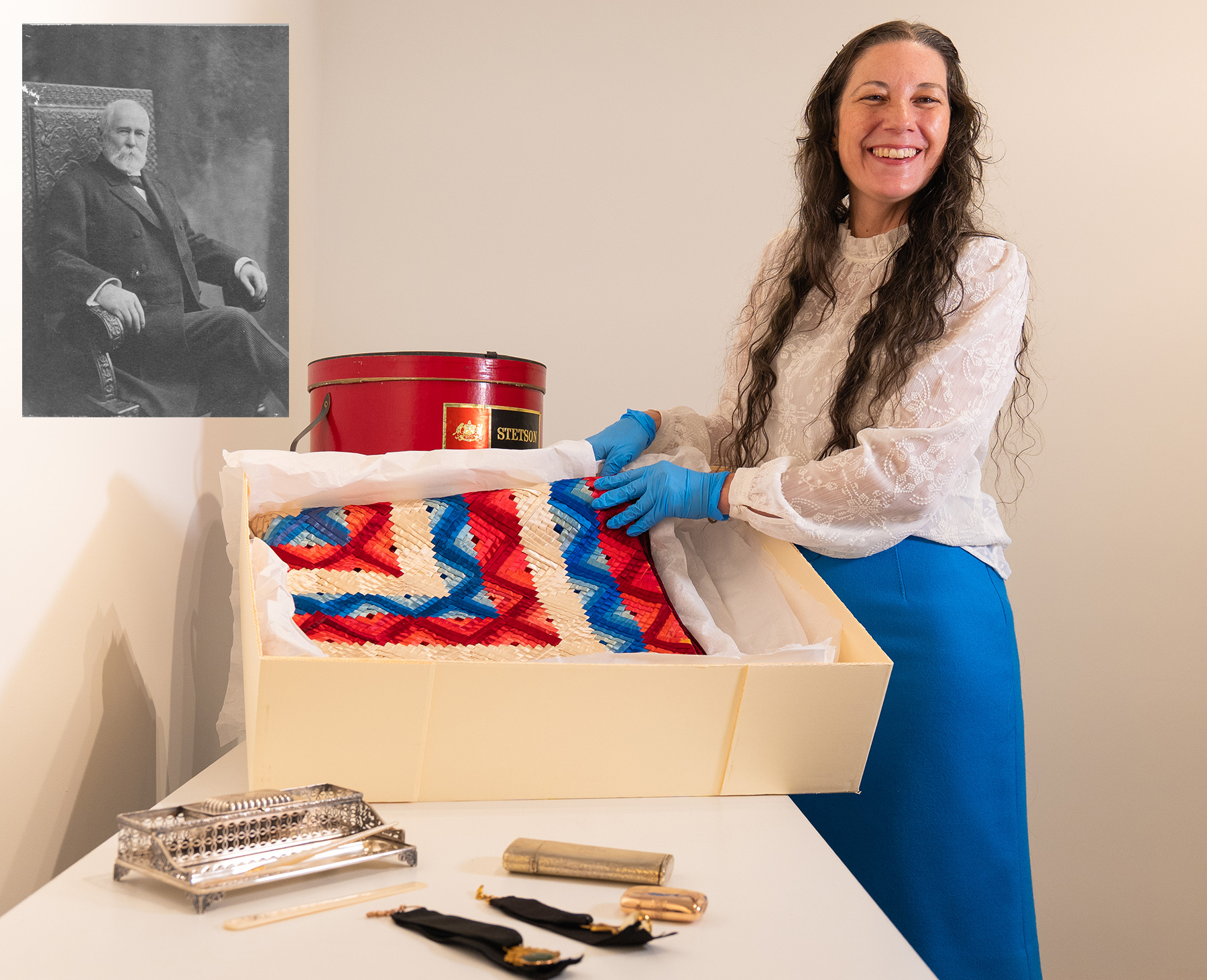 A woman stands with archival items, including a quilt, with a picture of John B. Stetson behind her.