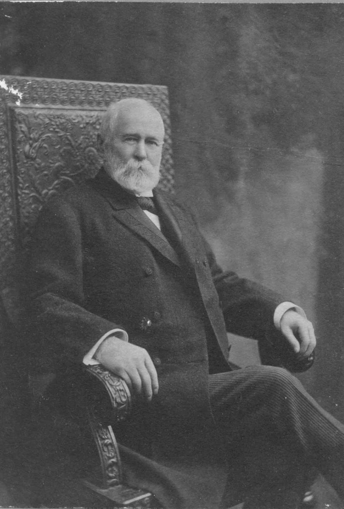 An older man sits in a high back arm chair in a grainy black and white photo.